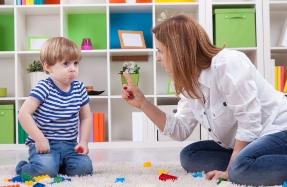 Mom and son on floor near toys and mom is scolding the son with finger pointing at him, he is a 2 year old