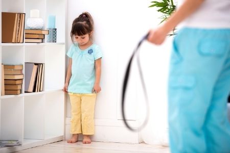 Stock image of child in corner looking down and out of focus adult holding looped belt