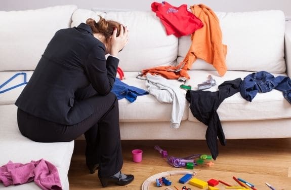 frustrated woman holding hands on head sitting in a very messy living room